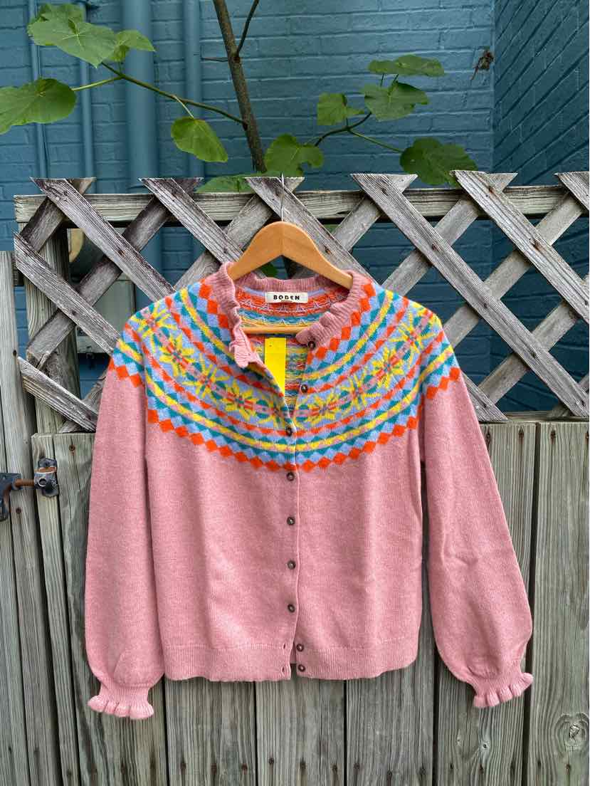 Pink cardigan with multicolored yoke pattern and button front, hanging on wooden hanger outdoors.
