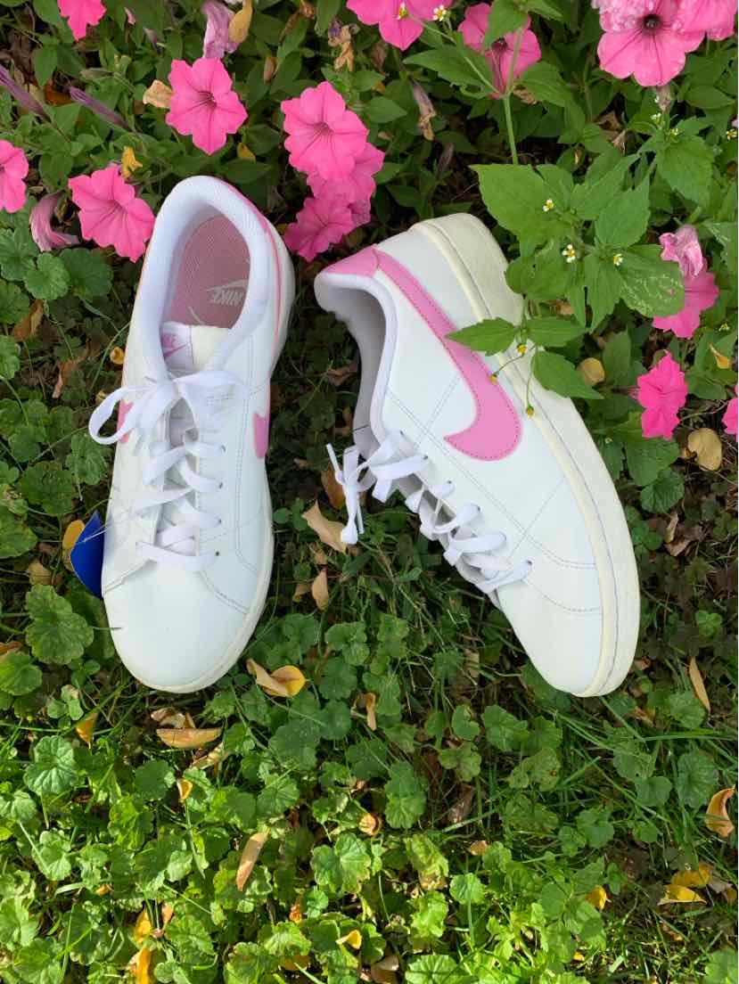 Pair of white Nike sneakers with pink swooshes and light gray mesh panels, resting on green foliage.