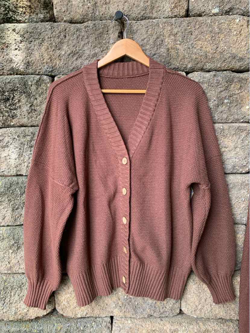 Brown knit cardigan with V-neck, button front, and ribbed cuffs and hem hanging on wooden hanger.