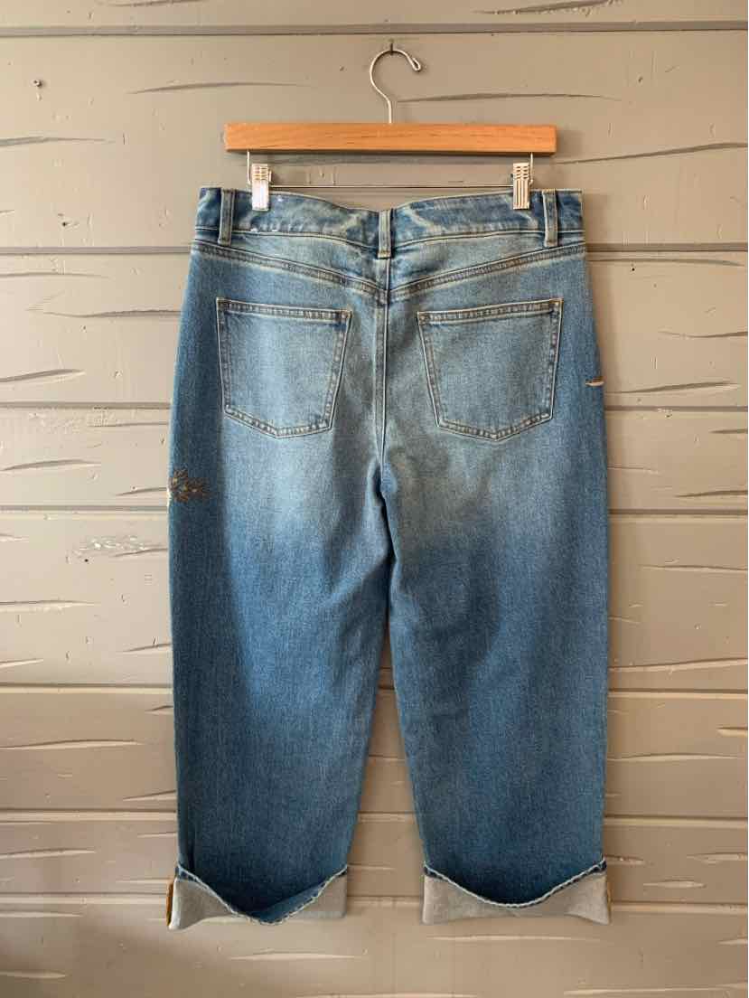 Light blue denim jeans with rolled cuffs and back pockets hanging on wooden hanger against wooden wall.
