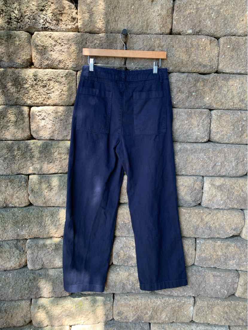 Navy blue straight-leg pants with back patch pockets hanging on wooden hanger against stone wall.