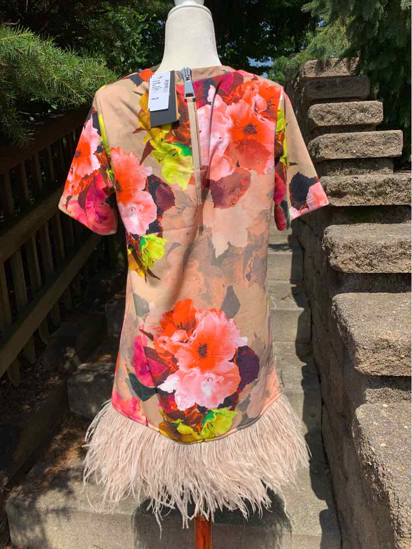 Short-sleeve floral dress with vibrant pink, orange, and green blooms, beige background, and feathered hem.