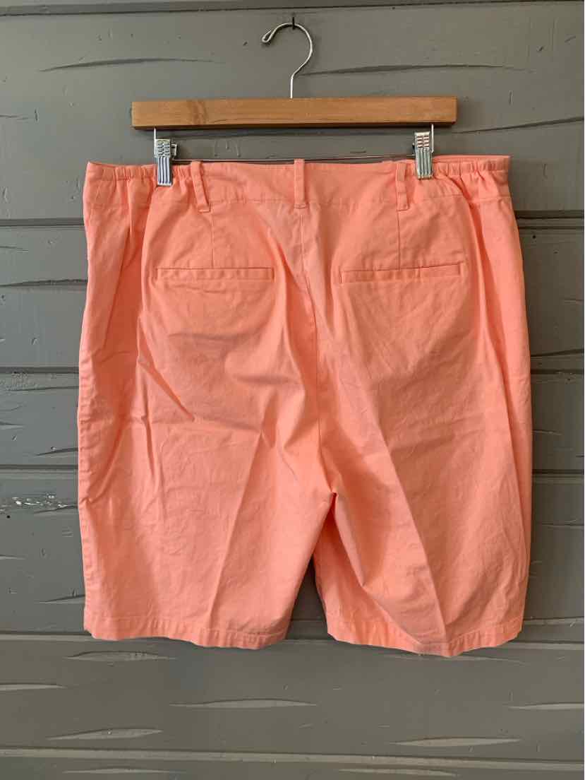Coral orange shorts with back patch pockets hanging on wooden hanger against gray wood wall.