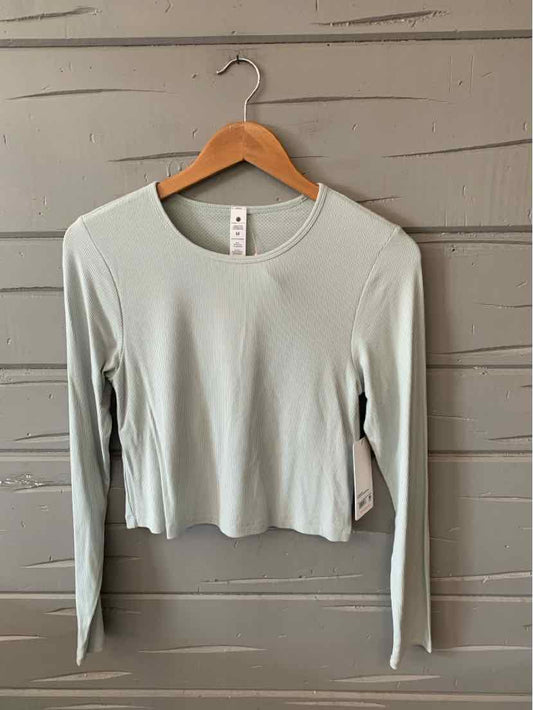 Mint green ribbed long-sleeve crop top with crew neckline hanging on wooden hanger against gray wooden wall.