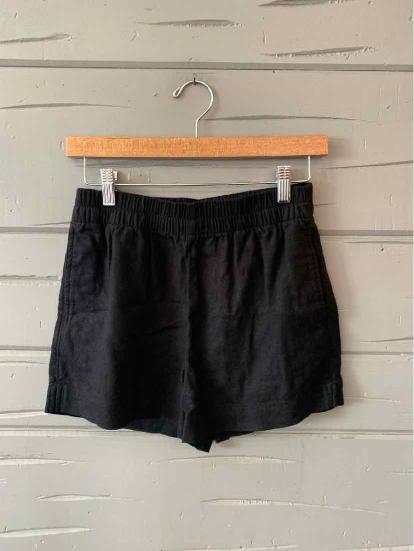 Black elastic waist casual shorts with side pockets hanging on wooden hanger against gray wall.