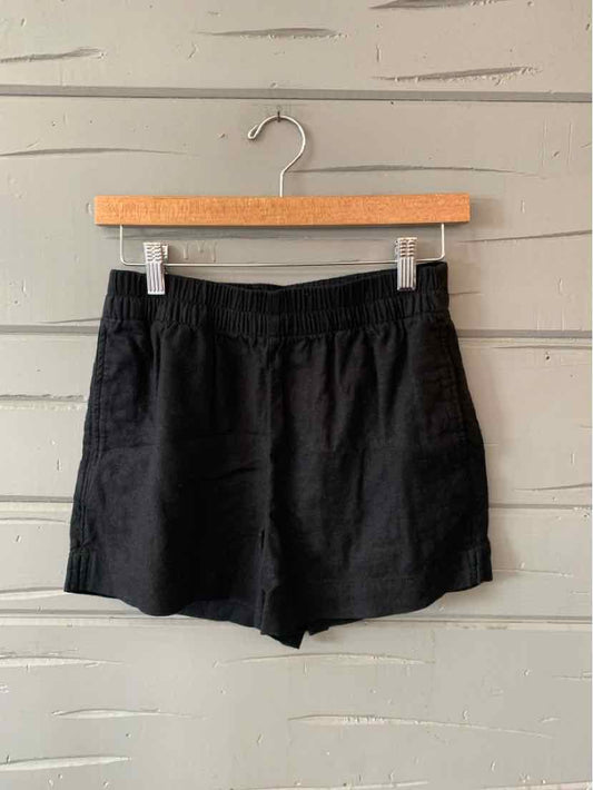 Black elastic waist casual shorts with side pockets hanging on wooden hanger against gray wall.