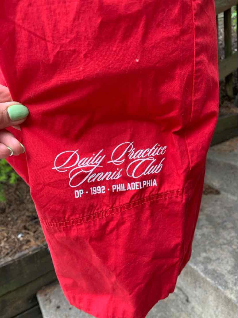 Red lightweight fabric pant leg with white "Daily Practice Tennis Club" text and logo embroidery.