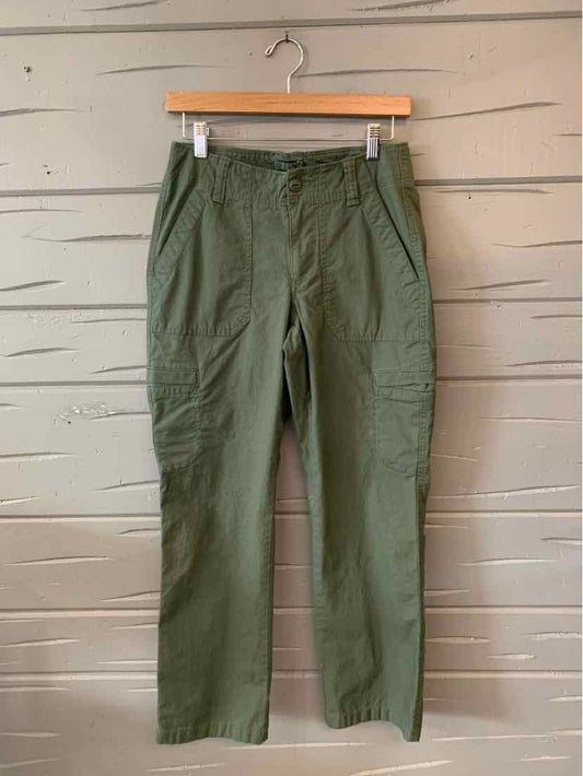 Olive green casual pants with straight legs and front pockets hanging on wooden hanger against gray wood wall.