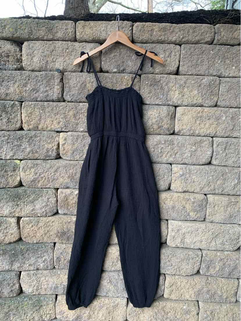 Black sleeveless jumpsuit with adjustable straps and gathered elastic waist hanging on wooden hanger.