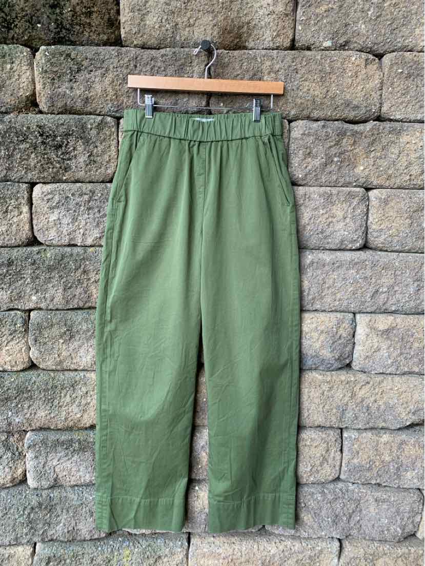 Olive green relaxed-fit pants with elastic waistband and straight legs hanging on wooden hanger against stone wall.