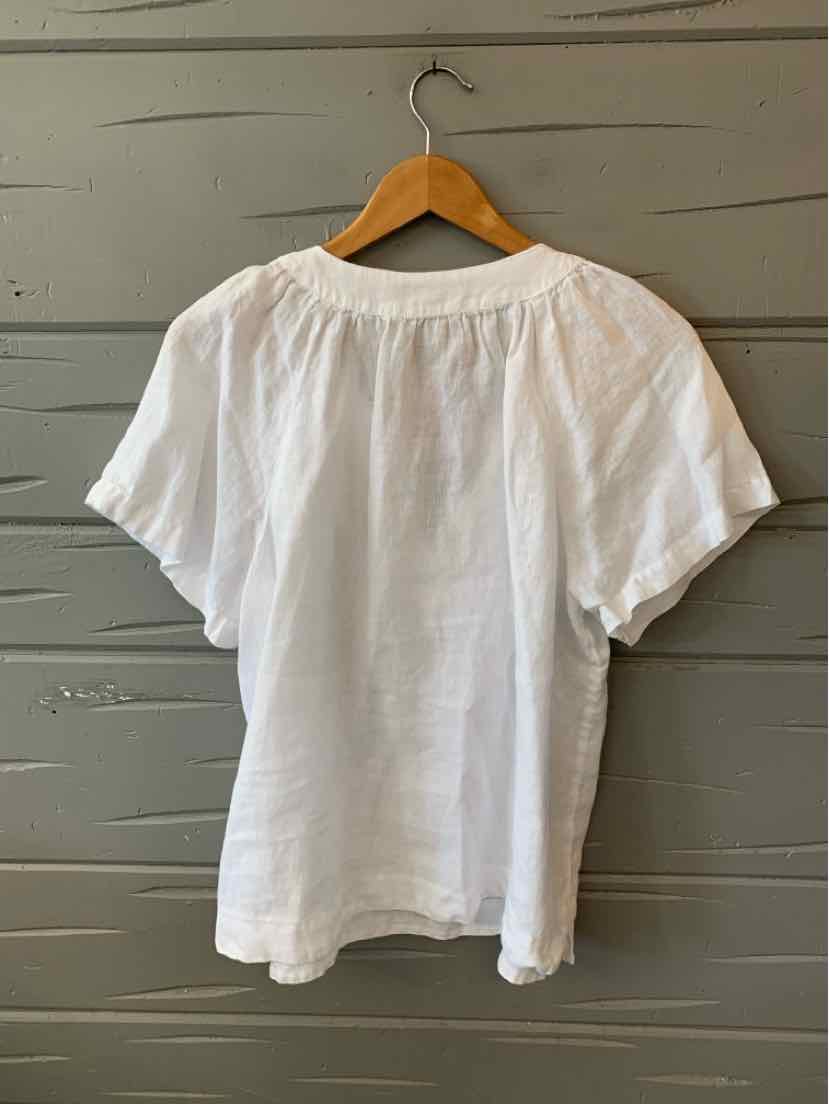 White short-sleeve blouse with round neckline and relaxed fit on wooden hanger against gray wall.