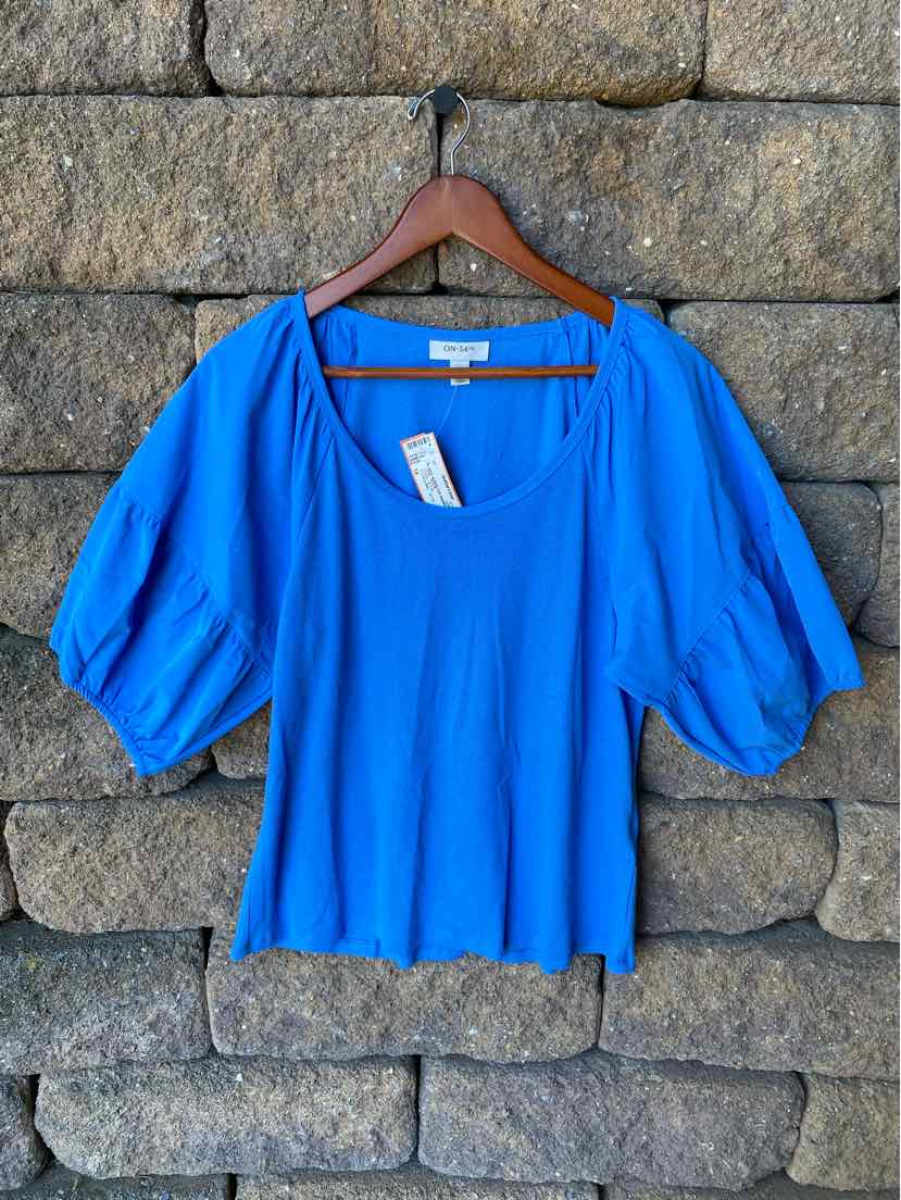 Bright blue blouse with puffed elbow-length sleeves and a scoop neckline, hanging on wooden hanger against stone wall.