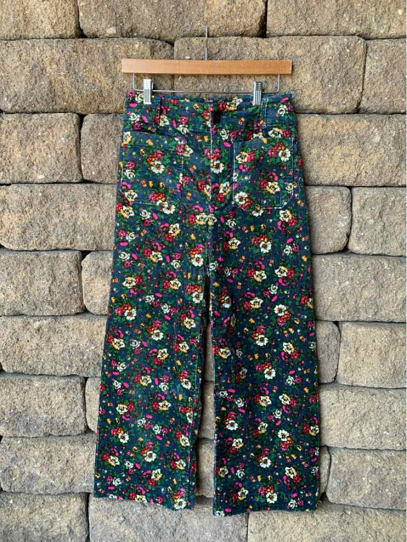 High-waisted dark green pants with vibrant floral print and wide-leg silhouette hanging on wooden hanger against stone wall.
