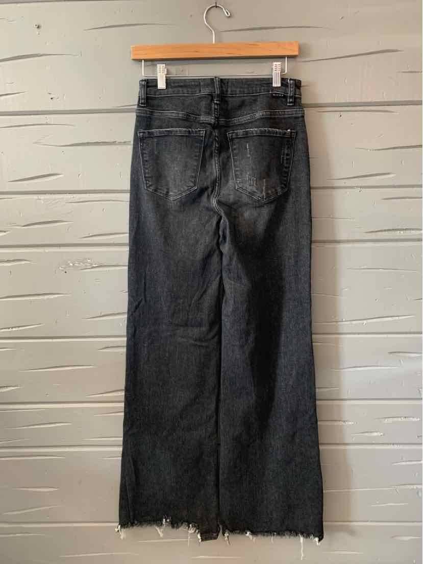 Black high-waisted denim jeans with wide legs and frayed hems hanging on wooden hanger against gray wooden wall.
