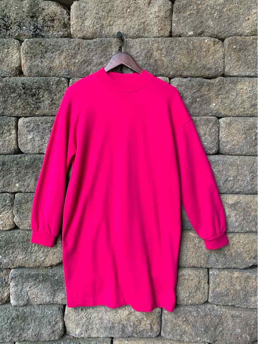 Bright pink long-sleeve dress with a mock neck and relaxed fit hanging against a stone wall background.