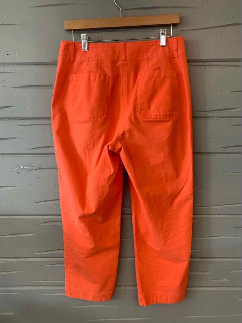 Bright orange cropped pants with back pockets hanging on wooden hanger against gray wall.