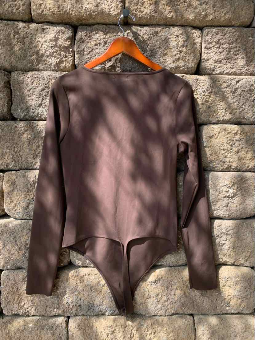 Long-sleeve fitted brown bodysuit with round neckline and snap closure bottom hanging on wooden hanger against stone wall.