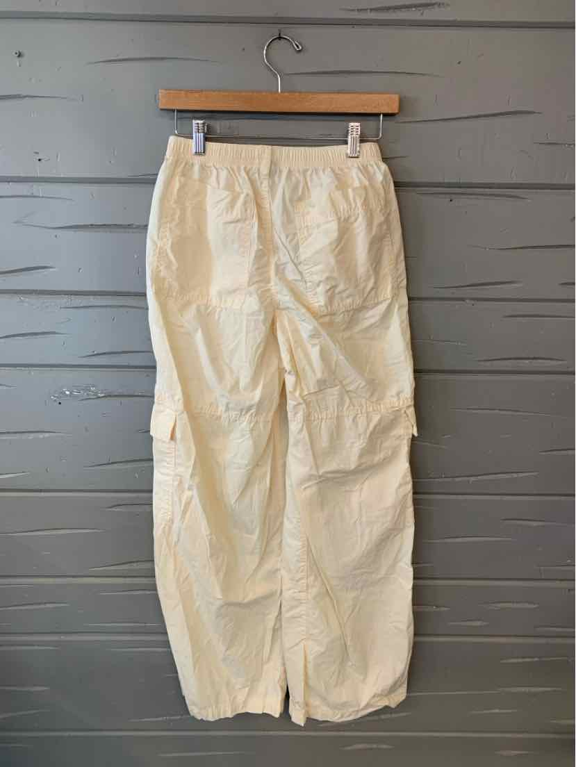 Cream-colored cargo pants with elastic waistband and multiple side pockets hanging on wooden hanger against gray wooden wall.