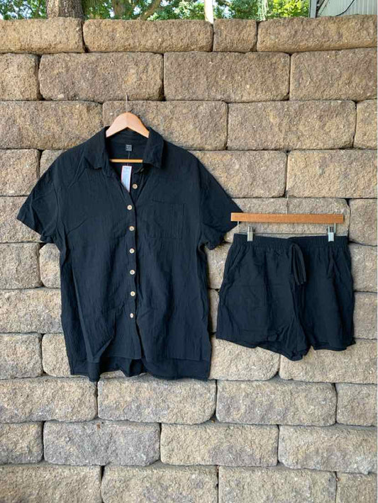 Black short-sleeve button-up shirt with matching elastic waist shorts hanging on wooden hangers against stone wall.