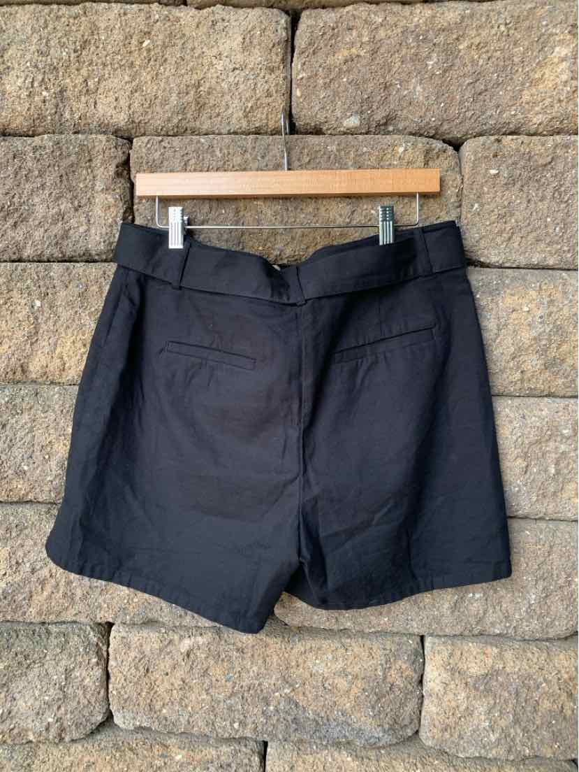 Black high-waist shorts with front pockets and belt loops hanging on wooden hanger against stone wall.