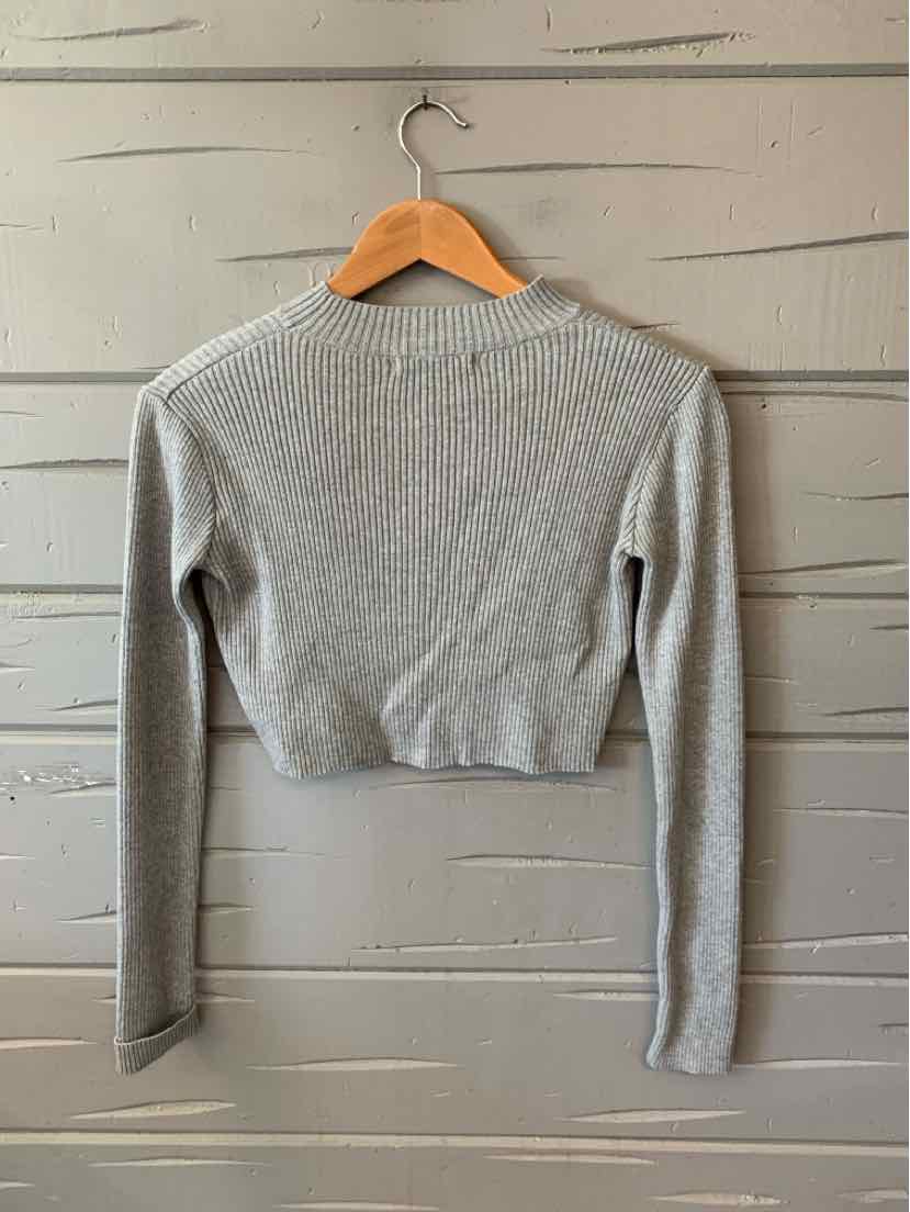 Light gray cropped knit sweater with long sleeves and ribbed cuffs on wooden hanger against gray wood panel background.