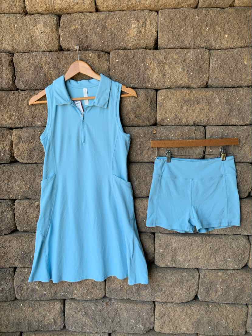 Light blue sleeveless hooded dress with front pockets and matching shorts on wooden hangers against a stone wall.