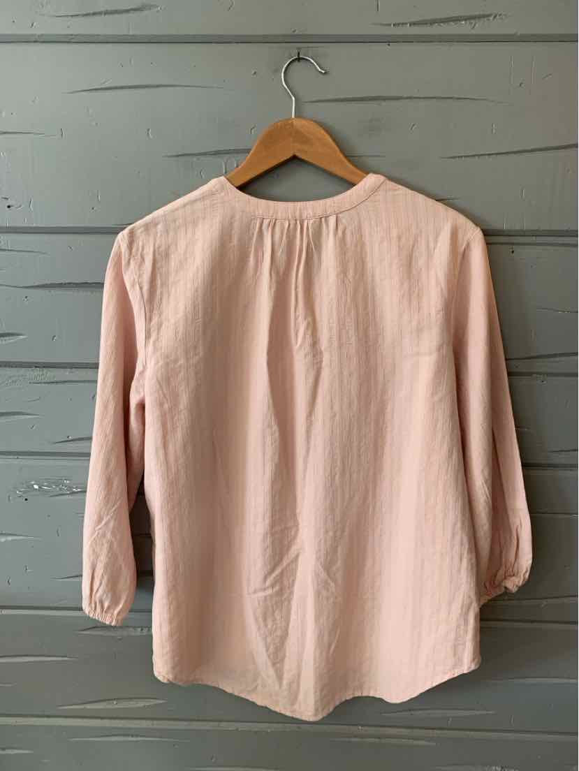 Light pink long-sleeve blouse with round neckline and buttoned cuffs hanging on wooden hanger against gray wooden wall.