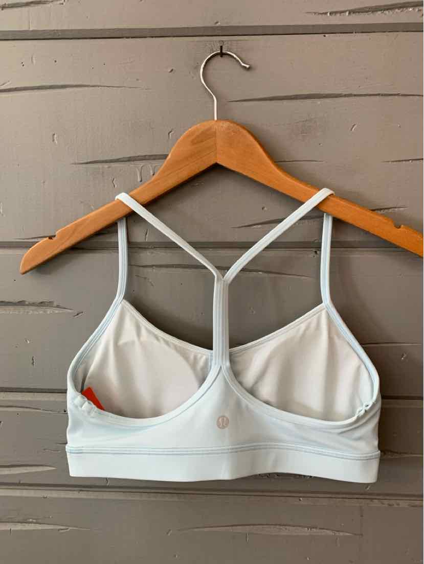 Light gray sports bra with thin straps and a racerback design on wooden hanger against gray wooden wall.