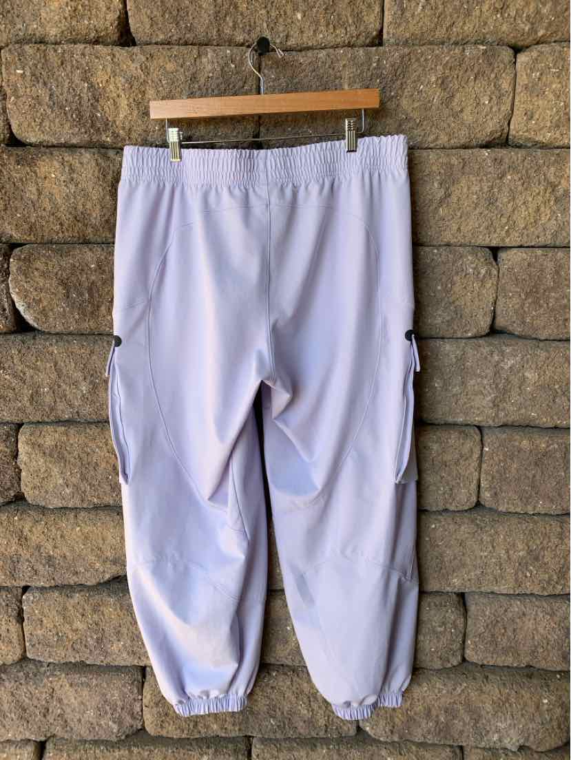 Light lavender Under Armour athletic jogger pants with an elastic waistband and gathered cuffs, hanging against a stone wall.