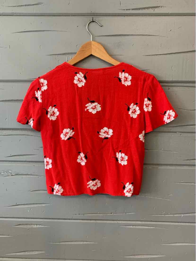 Red short-sleeve crop top with white floral print hanging on wooden hanger against gray wood background.