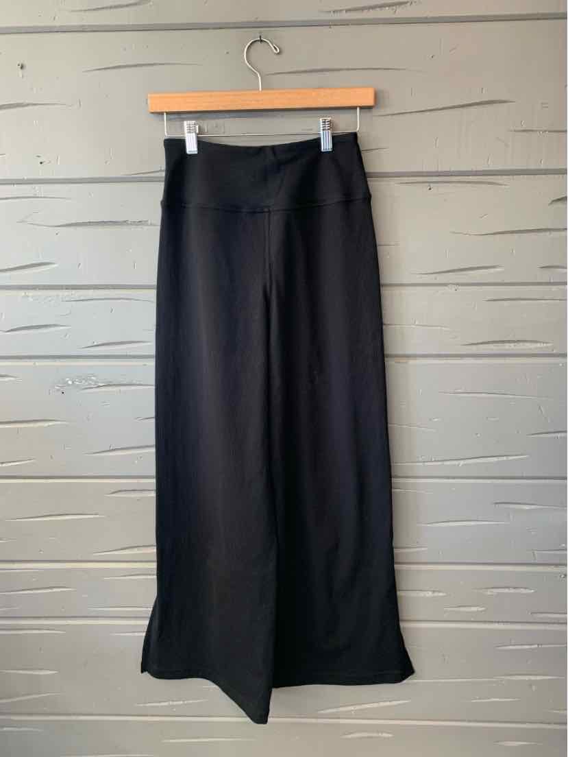Black Zella wide-leg trousers with a smooth finish, hanging from a wooden clip hanger.
