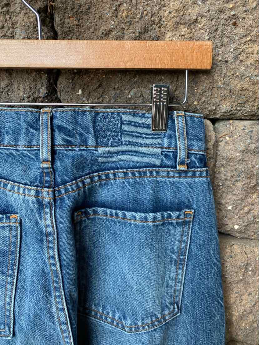 Blue denim jeans with classic back pockets and contrast stitching hanging on wooden hanger against stone background.