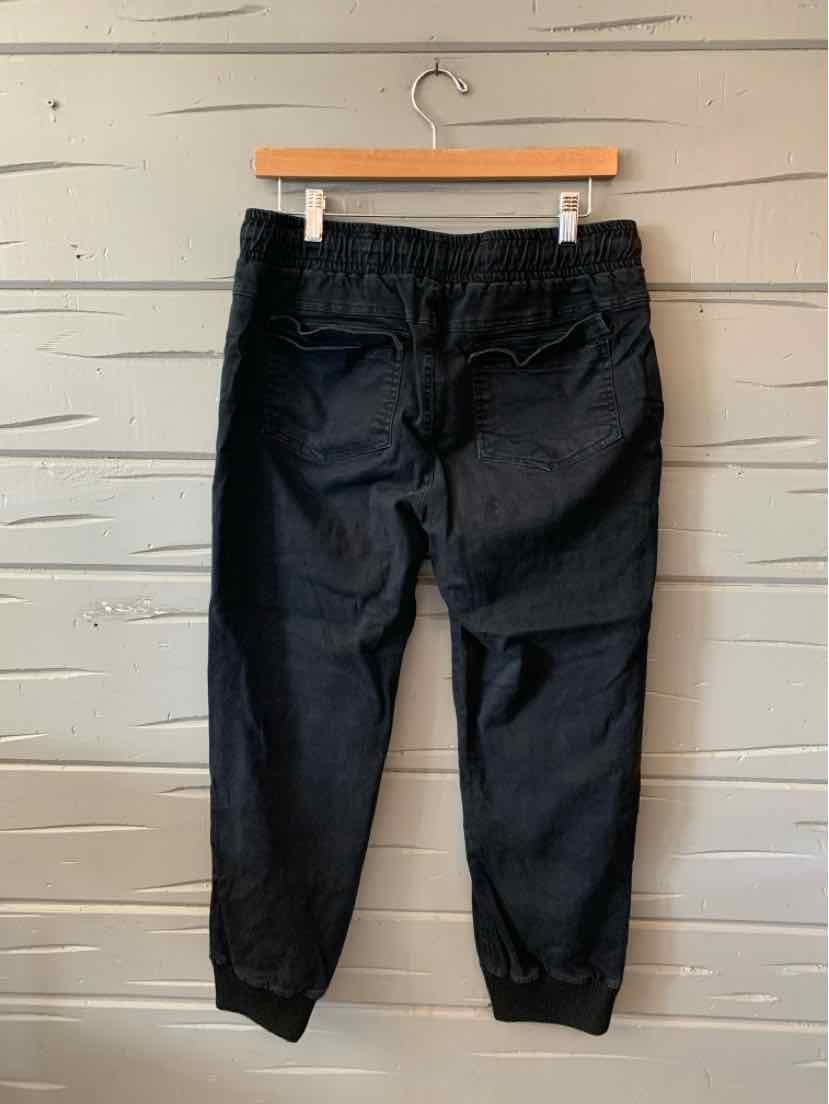 Black large solid pants with elastic waistband and cuffed ankles hanging on wooden hanger against textured gray wall.