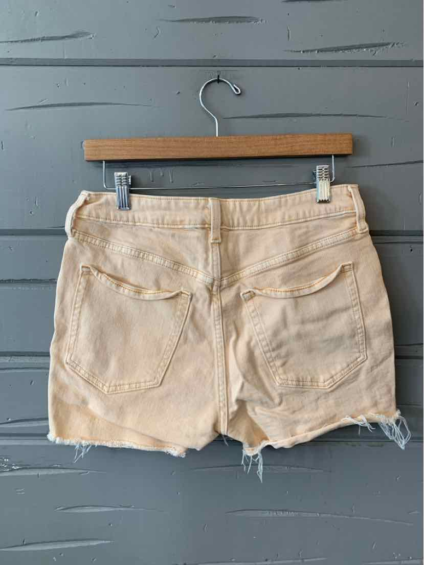 Light beige denim shorts with frayed hems and two back pockets hanging on wooden hanger against gray wall.