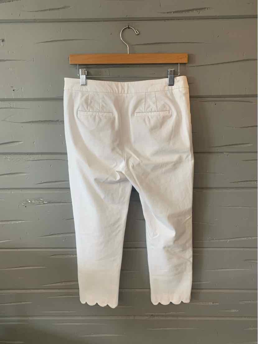 White cropped pants with scalloped hems and back pockets hanging on wooden hanger against gray wooden wall.