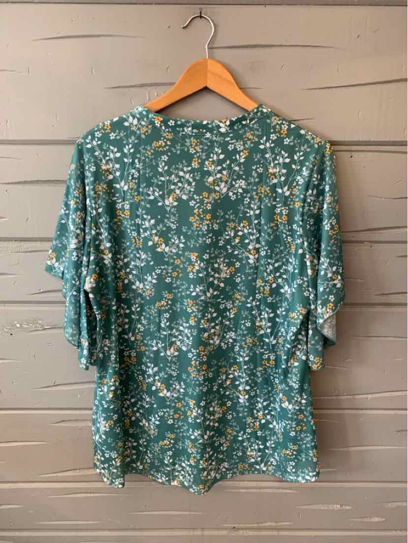 Green floral short-sleeve blouse with loose fit and round neckline hanging on wooden hanger.