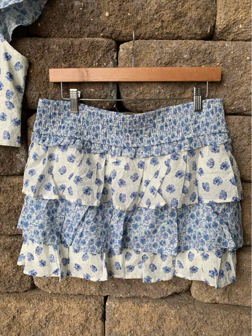 Blue and white paisley tiered mini skirt with elastic waistband hanging on wooden hanger against stone wall.