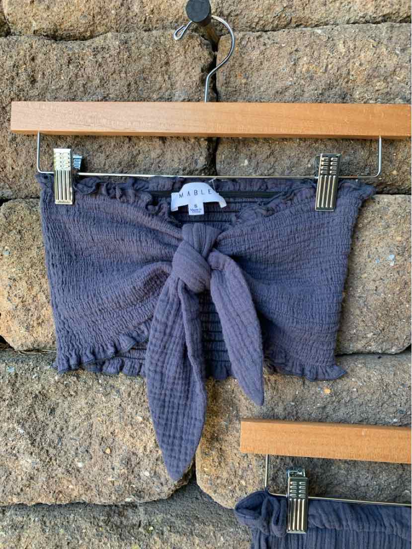 Dark gray textured crop top with front tie and pom-pom trim hanging on wooden hanger.