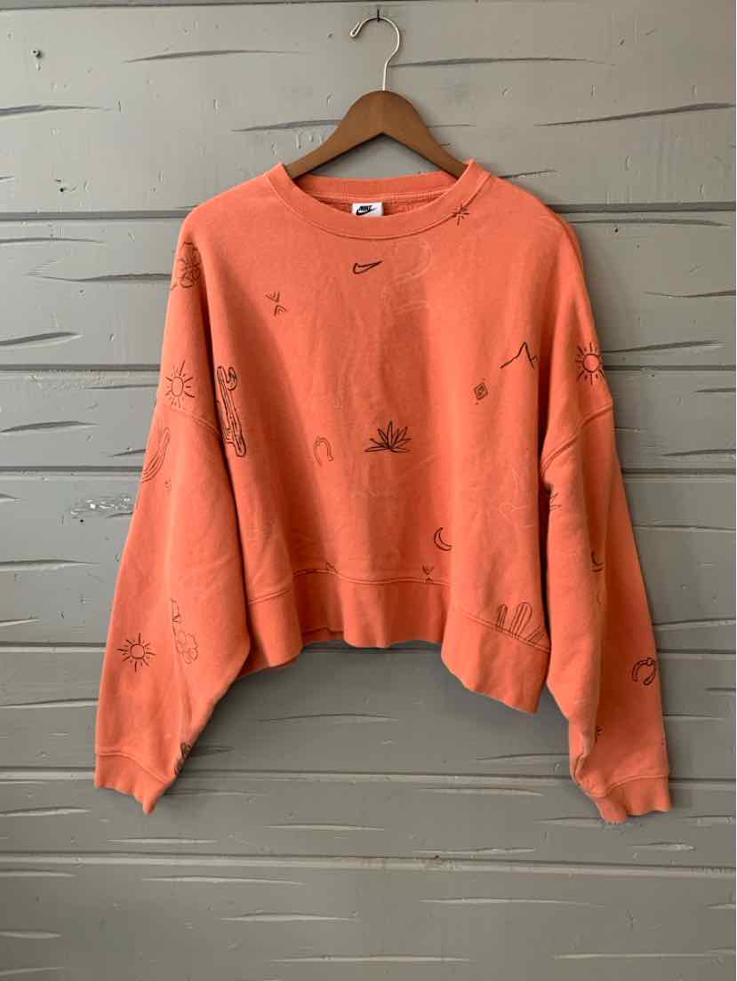 Coral orange cropped Nike sweatshirt with small black graphics and long sleeves on wooden hanger against gray wooden wall.