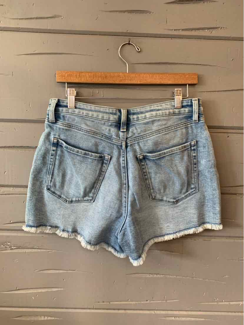 Light blue denim shorts with frayed hems and two back patch pockets hanging on wooden hanger against gray wall.