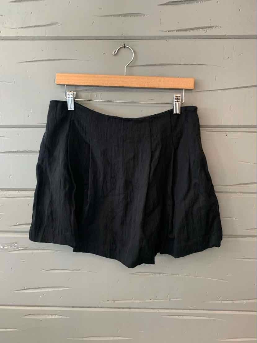 Black high-waist shorts with a loose fit and front pleats hanging on wooden hanger against gray slatted wall.