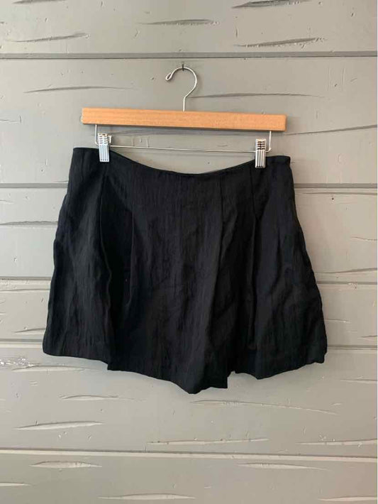 Black high-waist shorts with a loose fit and front pleats hanging on wooden hanger against gray slatted wall.