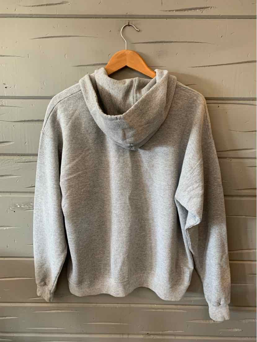 Light gray hoodie with a relaxed fit and a large hood hanging on a wooden hanger against a gray wooden wall.