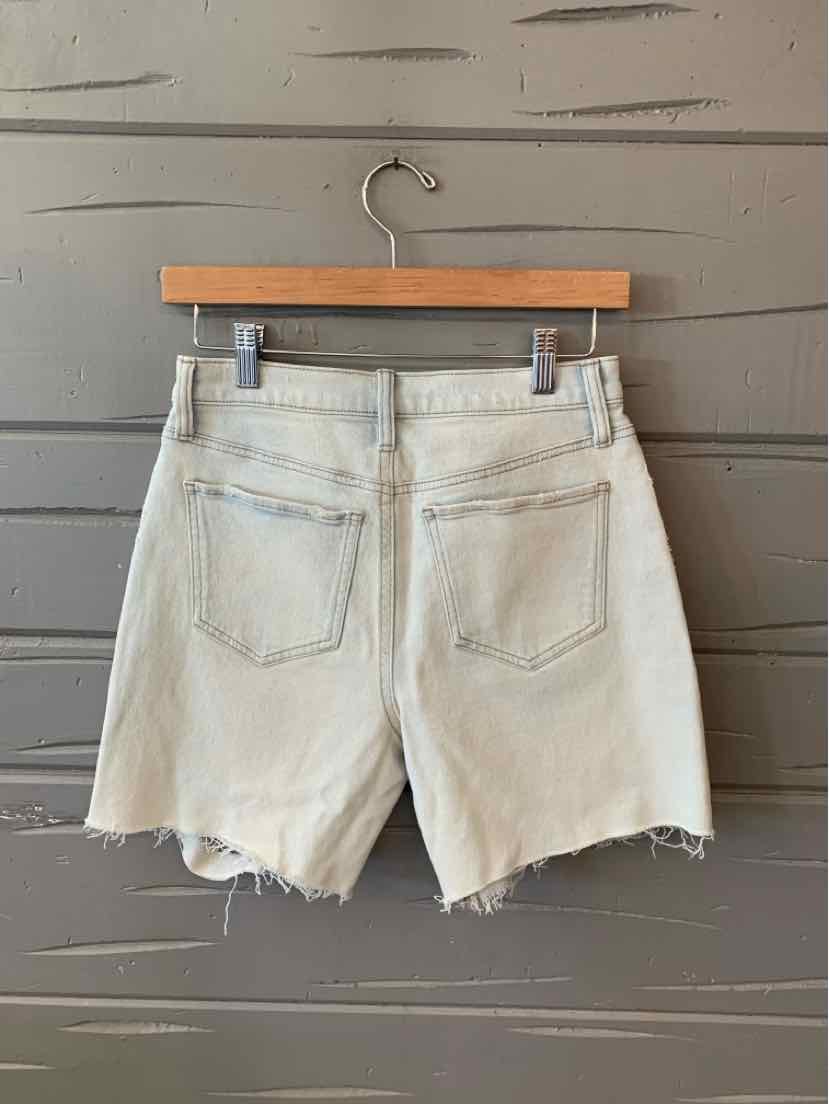 Light blue denim shorts with frayed hems and back pockets hanging on wooden hanger against gray wall.