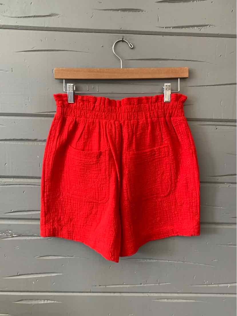 Bright red high-waisted shorts with a gathered elastic waistband and front patch pockets hanging on a wooden hanger.