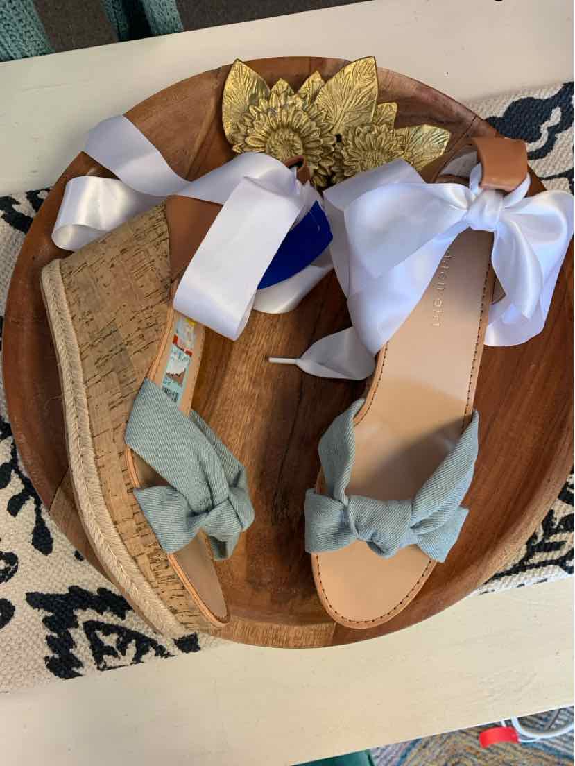 Pair of light blue flat sandals with knotted straps and a brown wedge sole displayed on a wooden tray.
