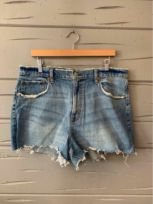 Blue denim shorts with frayed hems and front pockets hanging on wooden hanger against wood panel background.
