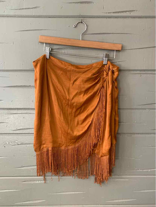 Bronze asymmetrical wrap skirt with side tie and fringe hem hanging on wooden hanger against gray wall.