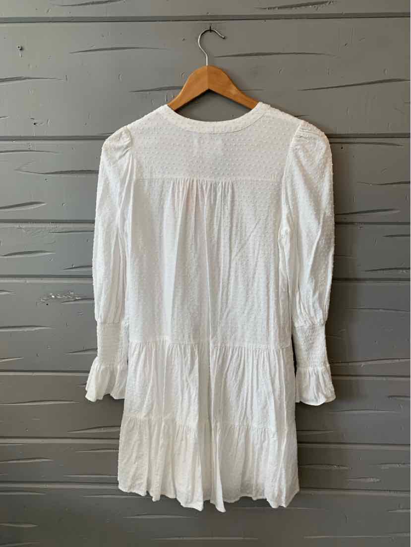 White long-sleeve dress with ruffled cuffs and tiered skirt hanging on wooden hanger against gray wall.