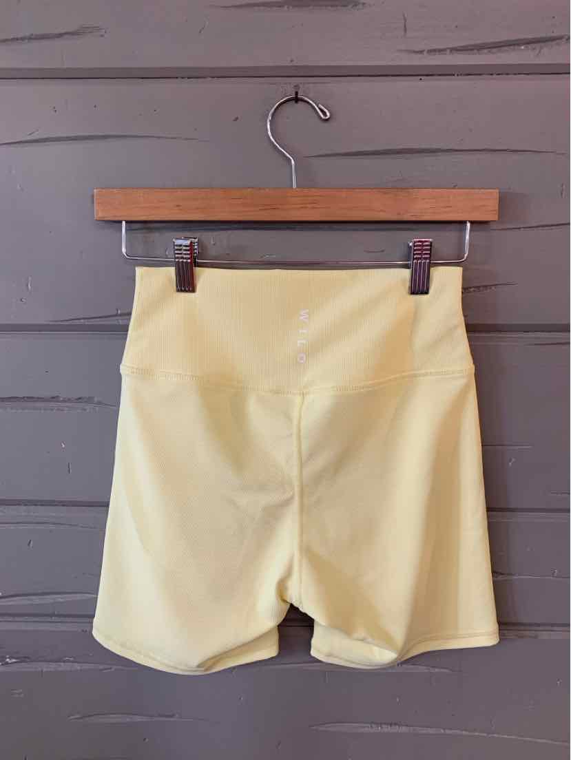 Light yellow high-waisted shorts with a smooth finish hanging on wooden hanger against gray wall.